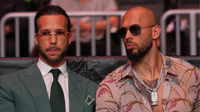 Two men, appearing to be Andrew Tate and his brother Tristan Tate, sit together at what looks like a combat sports event, with one wearing a teal suit and glasses and the other in a pink patterned shirt with sunglasses and a pearl chain necklace.