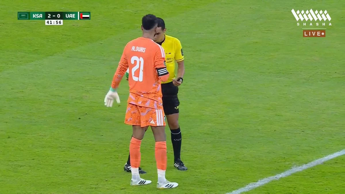 A Saudi goalkeeper named Kloivns, wearing the orange #21 jersey, stands on the pitch discussing with the yellow-shirted referee during the KSA vs UAE Gulf Cup final, with the scoreboard showing KSA leading 2-0 at 41:56, moments after the referee overturned a UAE penalty kick via VAR review.