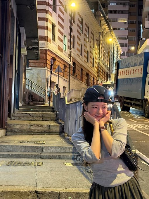 A nighttime street scene featuring a smiling young woman in a grey sweater and backwards cap posing on a sidewalk in Hong Kong.