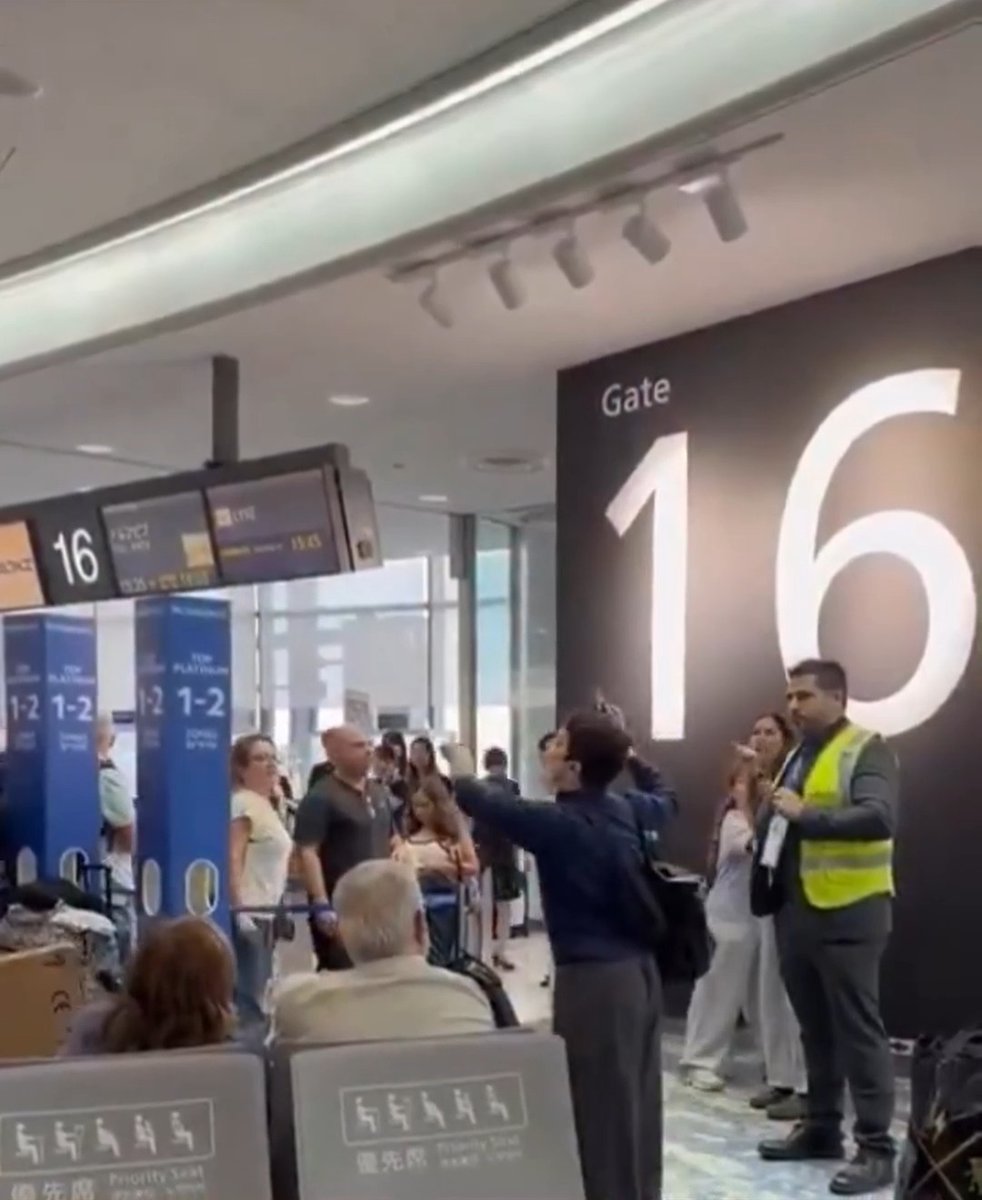 A group of passengers gathers at Gate 16 in an airport, with a man in a yellow safety vest and travelers raising their arms, appearing to sing or chant together. This image visually captures the moment described in the post, where Israeli travelers responded to a "Free Palestine" activist by singing "Am Israel Chai" ("The People of Israel Live"), demonstrating unity and resilience in the face of hostility.
