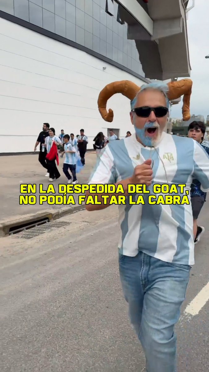 A passionate Argentine soccer fan dressed as a goat (cabra) — wearing ram horns, a blue-dyed beard, and sunglasses — runs joyfully outside what appears to be a stadium, likely River Plate's Monumental, in a light blue and white Argentina jersey. The Spanish text overlay reads "EN LA DESPEDIDA DEL GOAT, NO PODÍA FALTAR LA CABRA" (At the GOAT's farewell, the GOAT couldn't miss it), tying into the post describing the cabra's presence at Lionel Messi's sendoff match in Buenos Aires.
