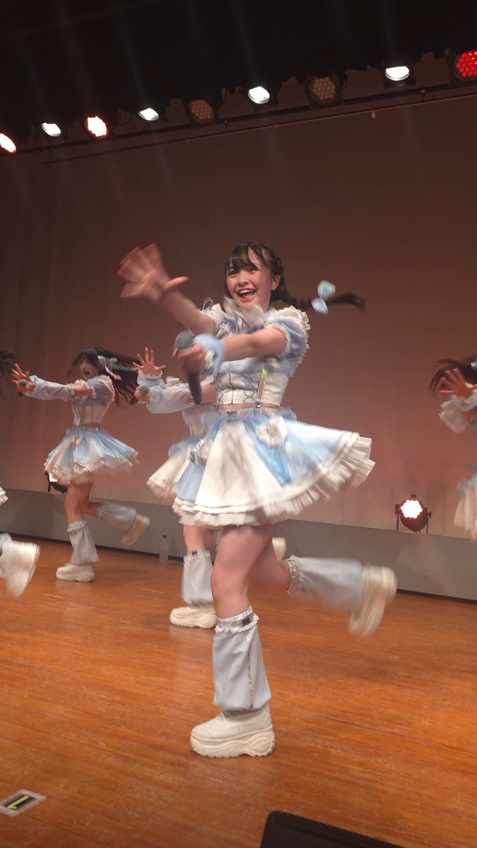 Young performers in light blue and white frilly costumes energetically dance on a wooden stage under bright lights.