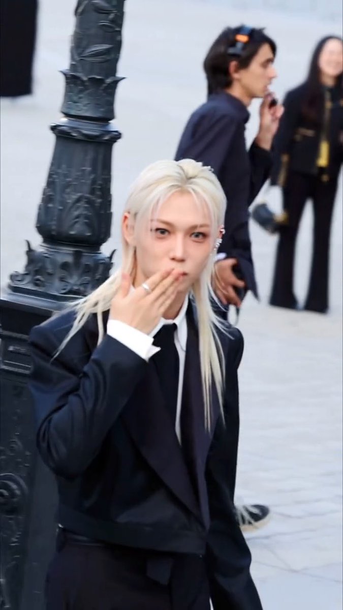 A blonde person wearing a black suit with a white shirt and black tie covers their mouth with one hand while posing outdoors near an ornate black metal lamppost.