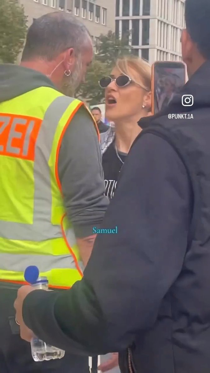 The photo shows a tense confrontation between a blonde woman wearing sunglasses and a police officer in a high-visibility "POLIZEI" vest, with bystanders recording the incident on their phones. Watermarks reading "@PUNKT.1A" and "Samuel" are visible on the image, consistent with the post's description of the scene as an altercation with Berlin police.