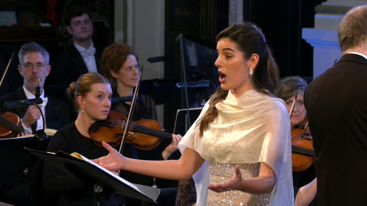 A female opera singer in a white shawl performs with an orchestra, singing with her mouth open while musicians play violins behind her.