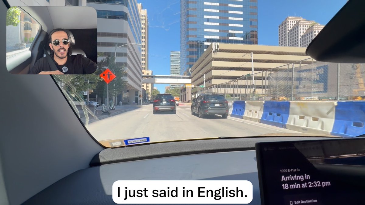 A dashcam view from inside a vehicle driving through a downtown city street, with an inset picture-in-picture showing a bearded man wearing sunglasses, accompanied by the caption 'I just said in English.' The dashboard display indicates arrival at 1000 E 41st St in 18 minutes at 2:32 pm.