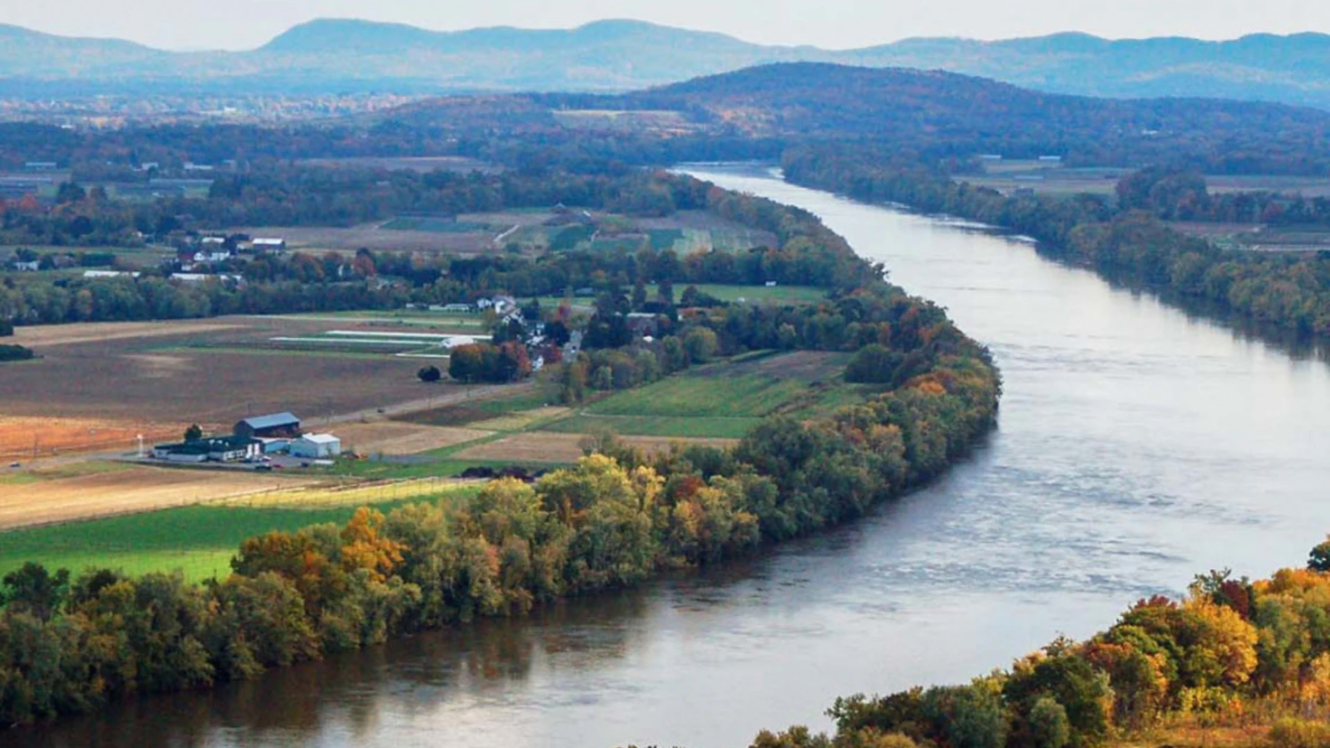 An aerial view of the Connecticut river winding through a patchwork of agricultural fields and trees with autumn foliage, with hills and mountains in the background, under a clear sky.