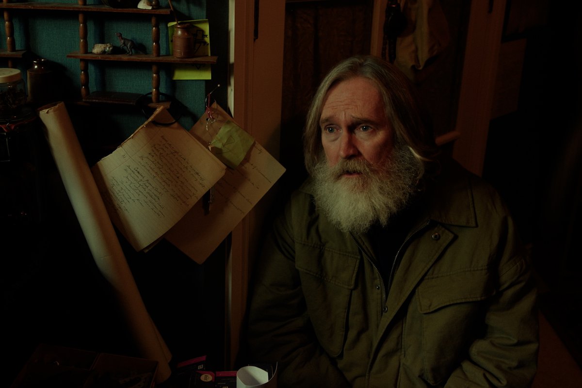 An older man with long gray hair and a thick beard sits in a dimly lit, cluttered room with handwritten notebooks hanging on the wall behind him. The image ties to the promotional post about "SmallProphets," evoking the show's mystical, alchemist-like atmosphere where fortune-telling and arcane rituals take center stage.