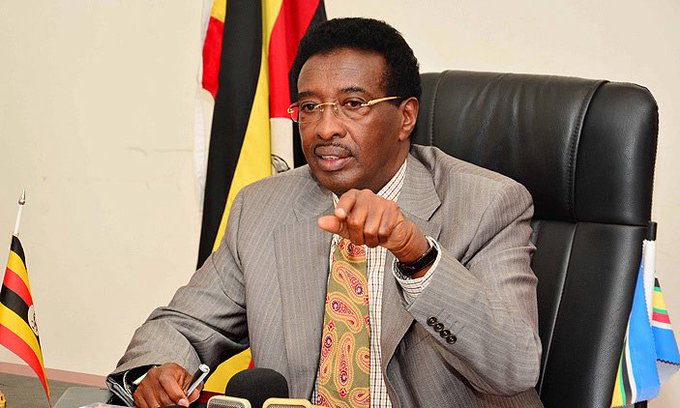 A man in a gray suit and patterned tie sits at a desk with microphones, gesturing with his hand while speaking, with Ugandan flags visible behind him. The caption relates to the post by suggesting the image depicts the official distancing himself from a controversy or scandal, presenting a contrast between his apparent public composure and the critical tone of the accompanying text.