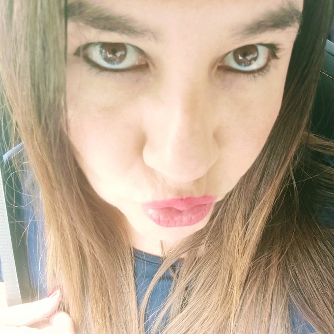 Close-up selfie of a young woman with long brown hair, striking light-colored eyes, and pink lipstick, looking directly at the camera.