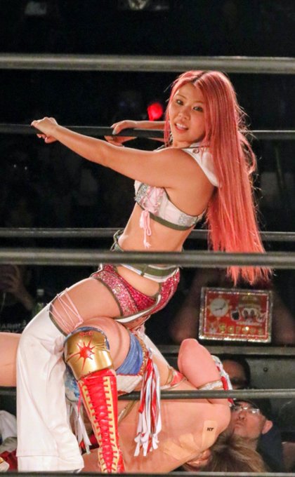 A female wrestler with long pink hair is performing a move on an opponent in a wrestling ring, wearing a pink and white ring outfit.