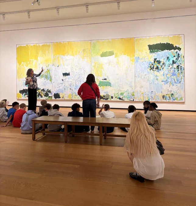 A group of students gathers in an art gallery, listening to a guide who points toward a large, multi-panel abstract painting featuring yellow, white, and green tones.