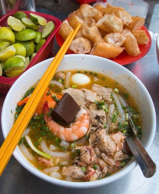A bowl of Vietnamese soup with shrimp, pork, quail egg, noodles, and herbs is accompanied by a side of fried dough sticks (youtiao) and a basket of limes.