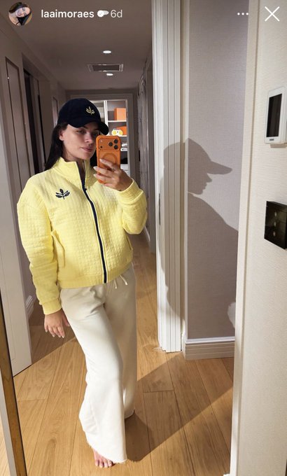 A woman takes a mirror selfie in a hallway, wearing a pale yellow zip-up Adidas jacket with the classic trefoil logo, white pants, and a black Adidas cap—showcasing Fenerium's sporty new design. The username "laaimoraes" is visible at the top of the Instagram story.