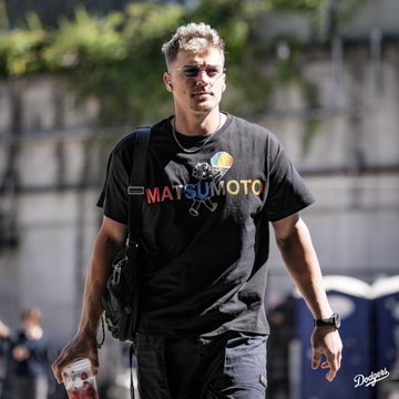 A young man with short blond hair walks outdoors wearing a black "MATSUMOTO" t-shirt and carrying a backpack and coffee cup, appearing to arrive at a stadium. The image likely relates to the #NLDS post, suggesting he may be a player arriving for the National League Division Series playoffs.