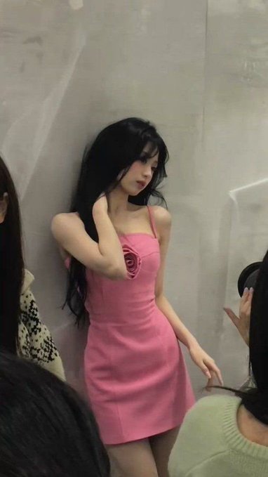 A photograph of a young woman with long black hair wearing a pink strapless dress, standing against a wall at what appears to be an indoor event.