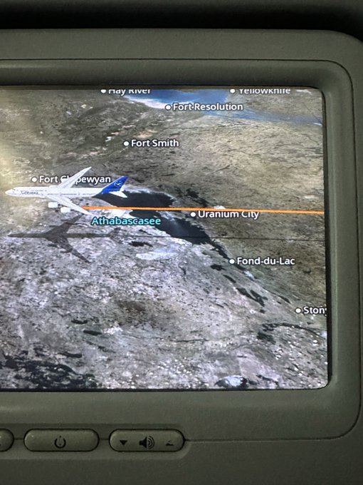 An in-flight entertainment screen displays a 3D map showing a Lufthansa airplane's flight path near Fort Chipewyan, Athabasca, and Uranium City over a lake and forest landscape.