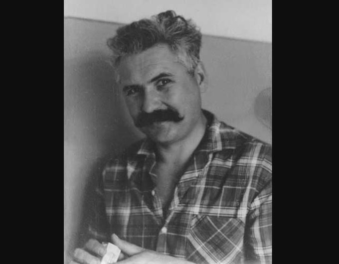 A black and white photograph of a middle-aged man with a mustache wearing a plaid shirt.