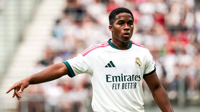 Action photograph of soccer player Vinícius Júnior wearing the white Real Madrid home kit with the 'Emirates FLY BETTER' sponsor logo during a match.