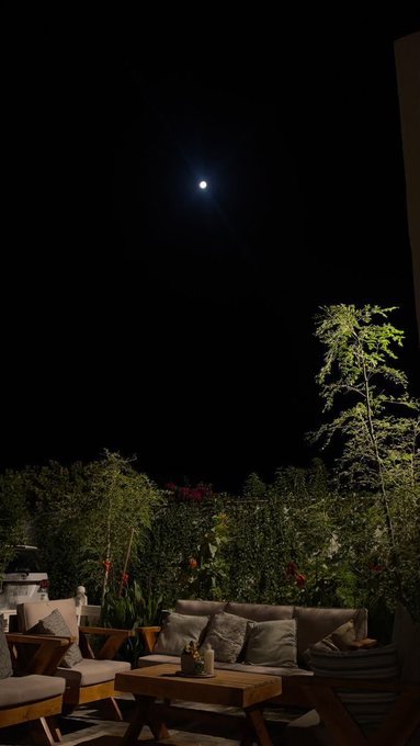 A nighttime outdoor scene showing a bright moon in a dark sky above a patio with outdoor furniture and garden plants.