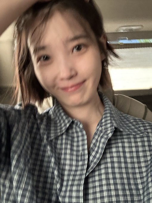A close-up selfie of a young woman with short brown hair wearing a blue and white plaid shirt, smiling softly at the camera.