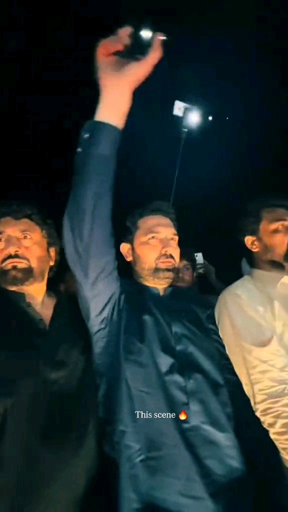 A man in a dark outfit holds up a smartphone with its flashlight on at night, with the caption 'This scene 🔥'.