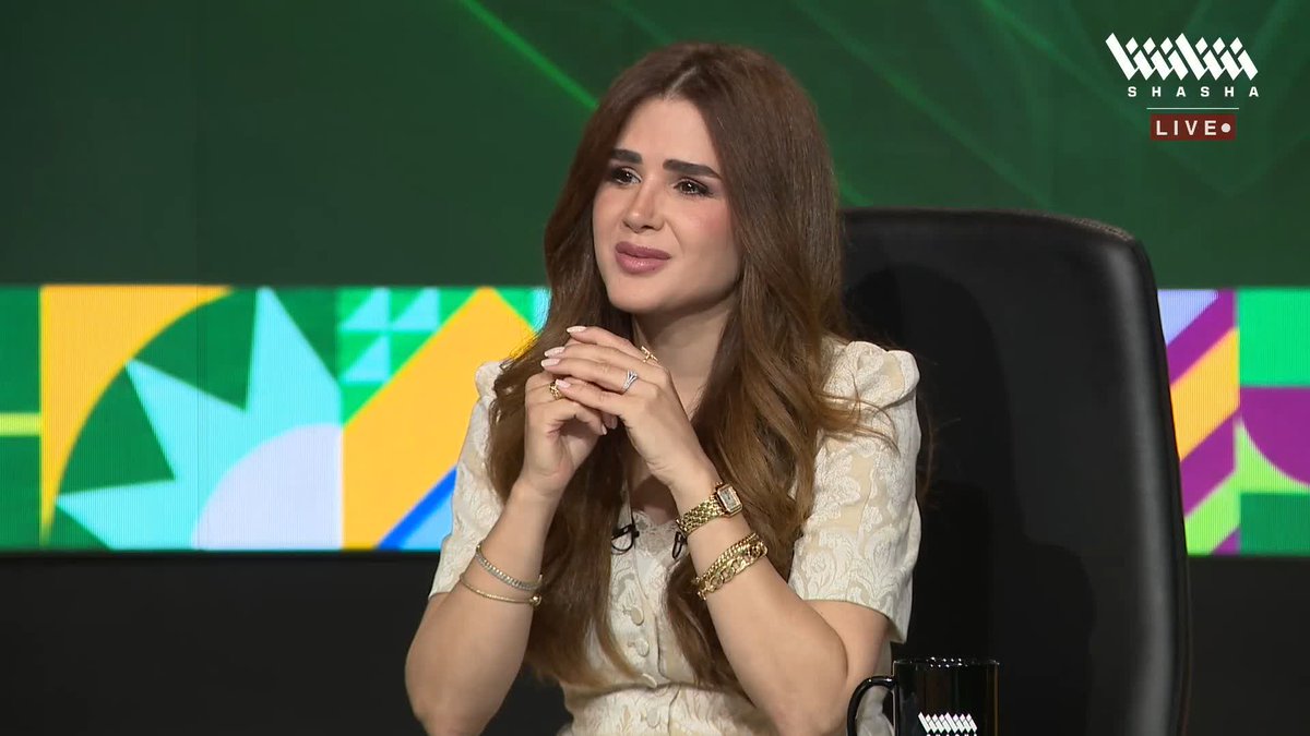 A woman in a cream embroidered dress sits at the Shasha Live broadcast desk, with her hands clasped thoughtfully near her face, during what appears to be a sports analysis segment. The colorful geometric backdrop and the post about congratulating Saudi people on the national team's revival suggest she is reacting to a match or commentary alongside host Mido.