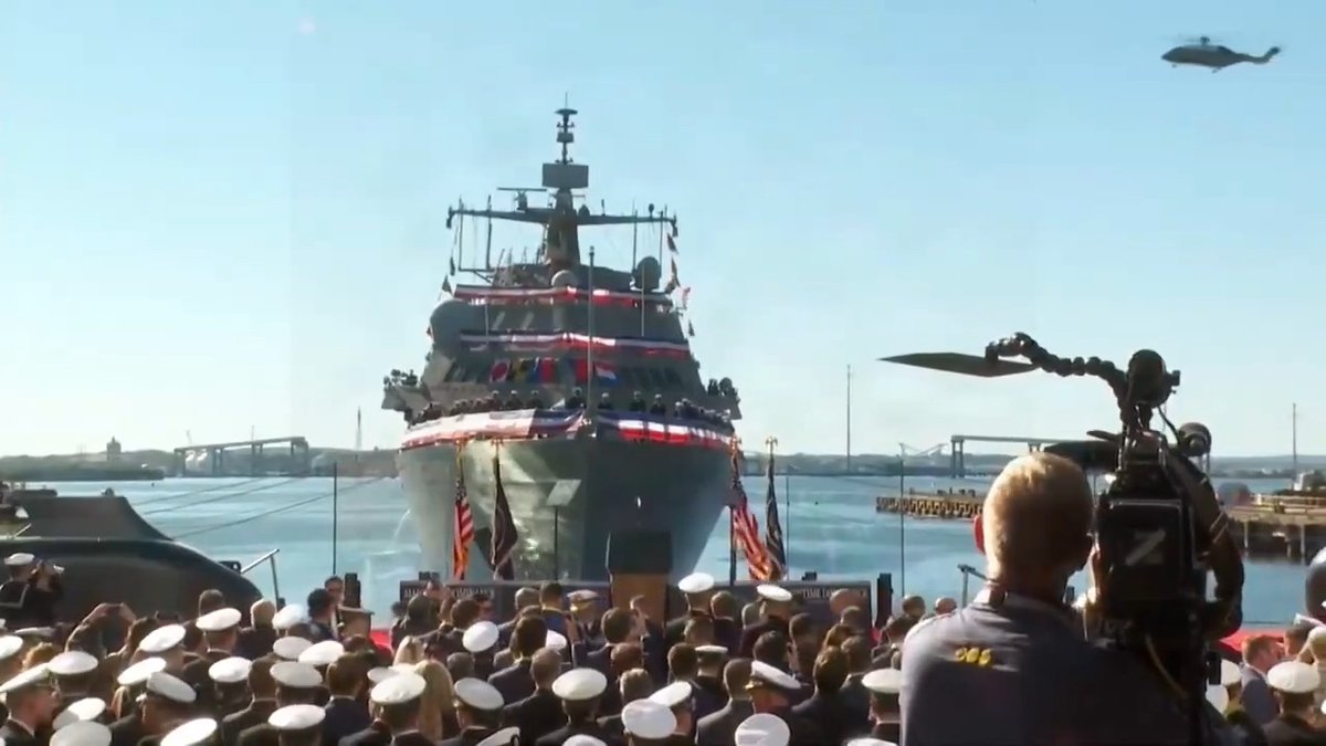 A military ship decorated with red, white, and blue bunting at what appears to be a commissioning ceremony, with sailors in white caps, a helicopter flying overhead, and a cameraman filming in the foreground.