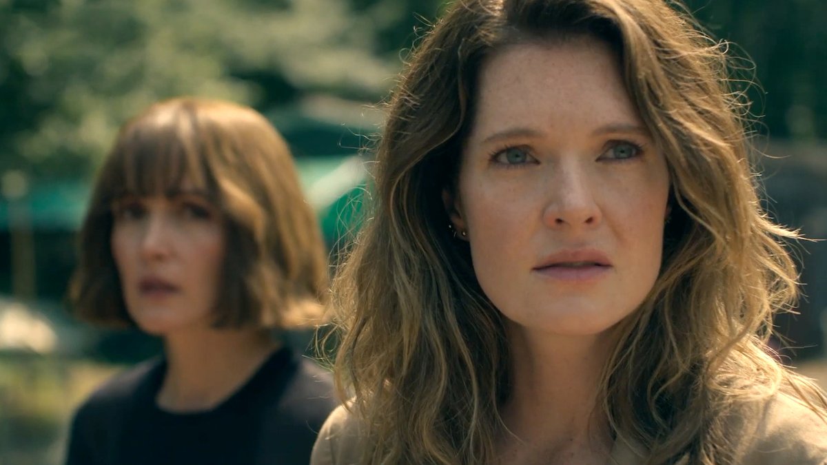 Two women stand outdoors with tense, focused expressions, the woman in the foreground featuring wavy auburn hair and freckles, while her companion with a sharp bob stands slightly behind. The image captures a dramatic moment from the Peacock series "TheGoodDaughter," starring Rose Byrne and Meghann Fahy, premiering November 12.