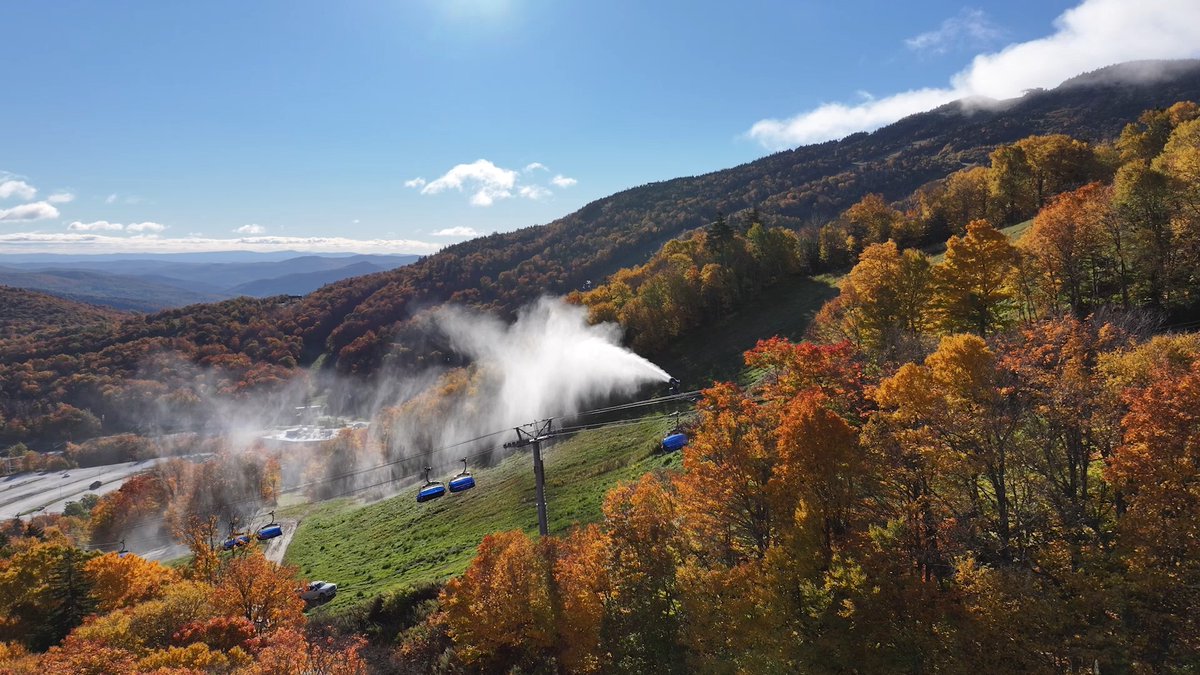 A scenic mountain landscape in autumn features colorful fall foliage, ski lift gondolas, and snow-making machines spraying artificial snow on a ski slope.
