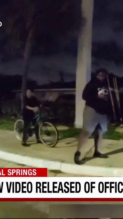 This image appears to be a low-quality, zoomed-in screenshot from surveillance or cellphone video showing a person on a bicycle and another individual standing nearby at night on what looks like a sidewalk, with a news chyron at the bottom reading "...AL SPRINGS" and "...VIDEO RELEASED OF OFFIC..." relating to the released footage of the fatal crash involving a police officer in Coral Springs.