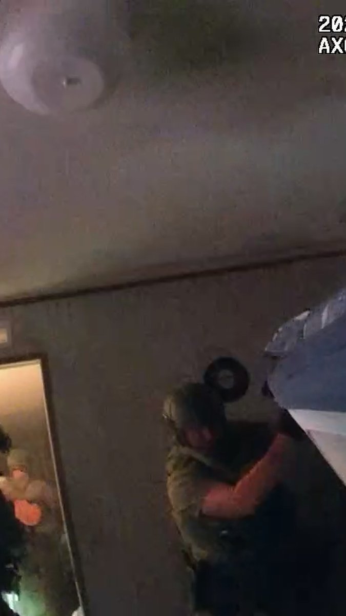A blurry image showing what appears to be a person wearing a helmet and gas mask holding a blue object, with a ceiling fan visible at the top.