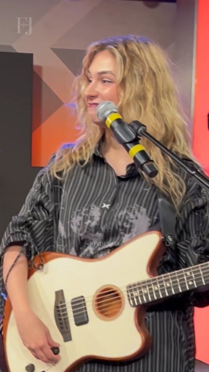 A blonde woman wearing a dark striped shirt smiles while playing a cream-colored acoustic guitar and singing into a microphone.
