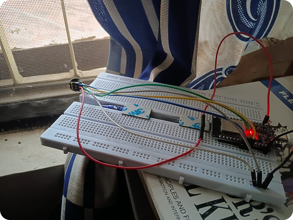 An electronics breadboard setup on a windowsill with various jumper wires connecting components, including a microcontroller with a glowing red LED, placed on top of a cardboard box.