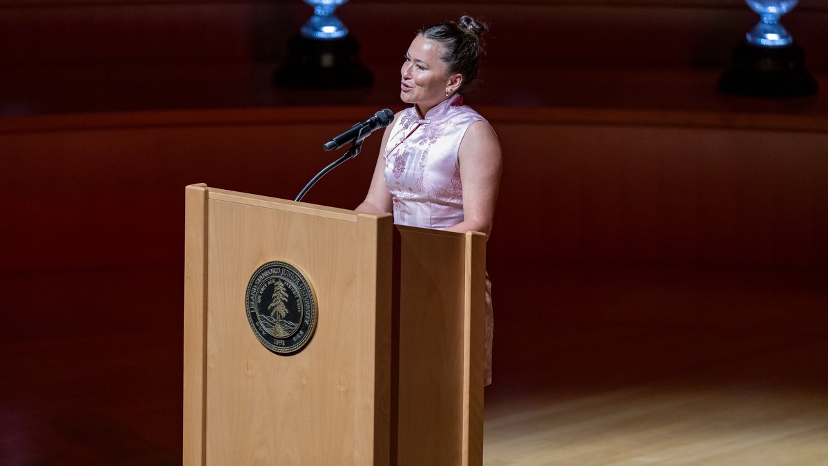 A woman in a pink patterned cheongsam-style dress stands at a wooden podium with the Stanford Junior University seal, speaking into a microphone.