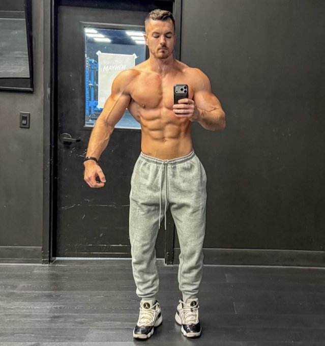 A muscular shirtless man takes a mirror selfie in a gym, flexing his well-defined abs and arms while wearing gray sweatpants and white sneakers. The image visually reinforces the post's muscle-building tip, showcasing a lean, defined physique as proof of effective high-carb, moderate-protein, low-fat nutrition. A partially visible sign in the background reads "MAYHEM," likely referring to the Mayhem Athlete brand associated with bodybuilding and fitness training.
