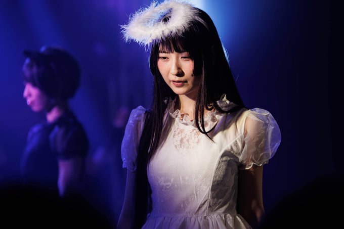 A young woman with long dark hair wearing a white frilly dress and a feathered headpiece stands on stage under moody blue lighting.