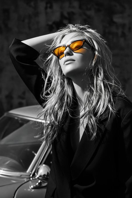 A black and white fashion-style portrait of a woman wearing a black blazer and orange-tinted sunglasses, standing next to a classic car.