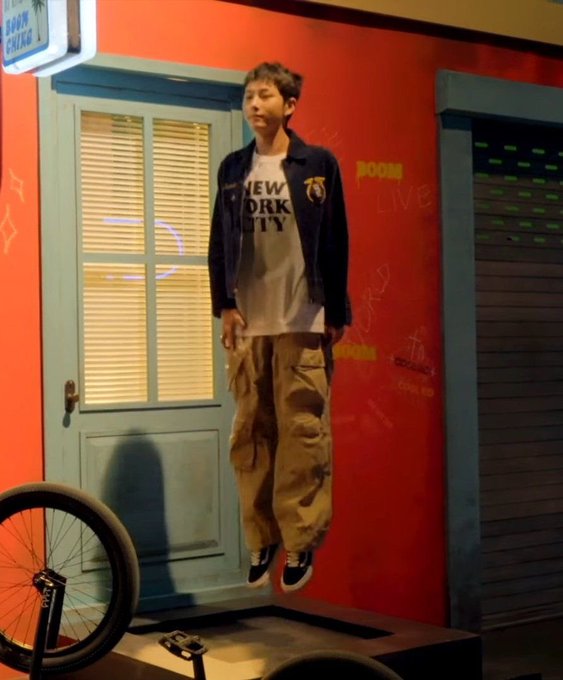 A young man appears to be jumping or levitating in front of a red wall with a 'BOOM LIVE' neon sign, wearing a 'New York City' t-shirt, denim jacket, and tan cargo pants, with a bicycle wheel visible in the foreground.