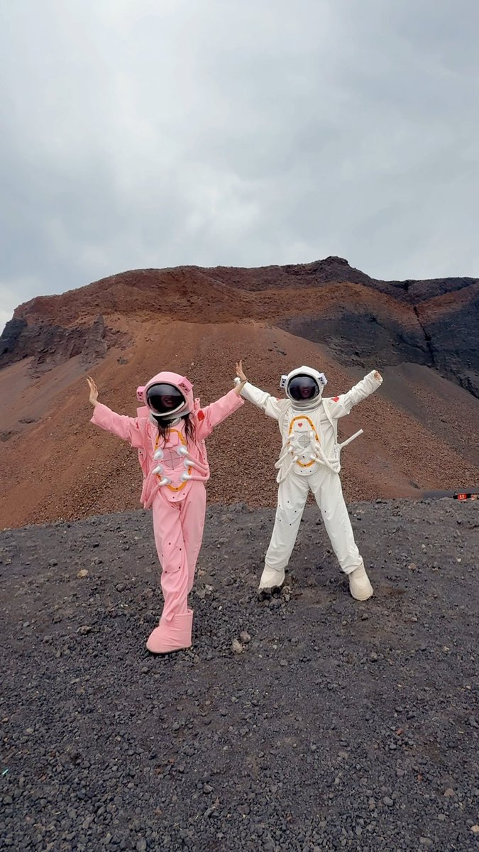Two people wearing pink and white astronaut costumes pose with arms raised on a rocky volcanic landscape under an overcast sky.