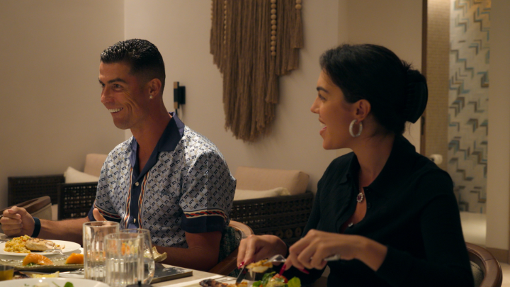 Cristiano Ronaldo and Georgina Rodríguez sit at a dinner table, with Ronaldo smiling broadly and Georgina laughing while cutting food on her plate, seemingly enjoying a meal together in a warmly lit room featuring decorative wall hangings. The image of Ronaldo's wide, eager smile aligns with the post's critique of his "arrogance" and need for validation, as he beams with apparent pleasure in a candid moment that contrasts with Ibrahimović's reportedly more self-assured demeanor.