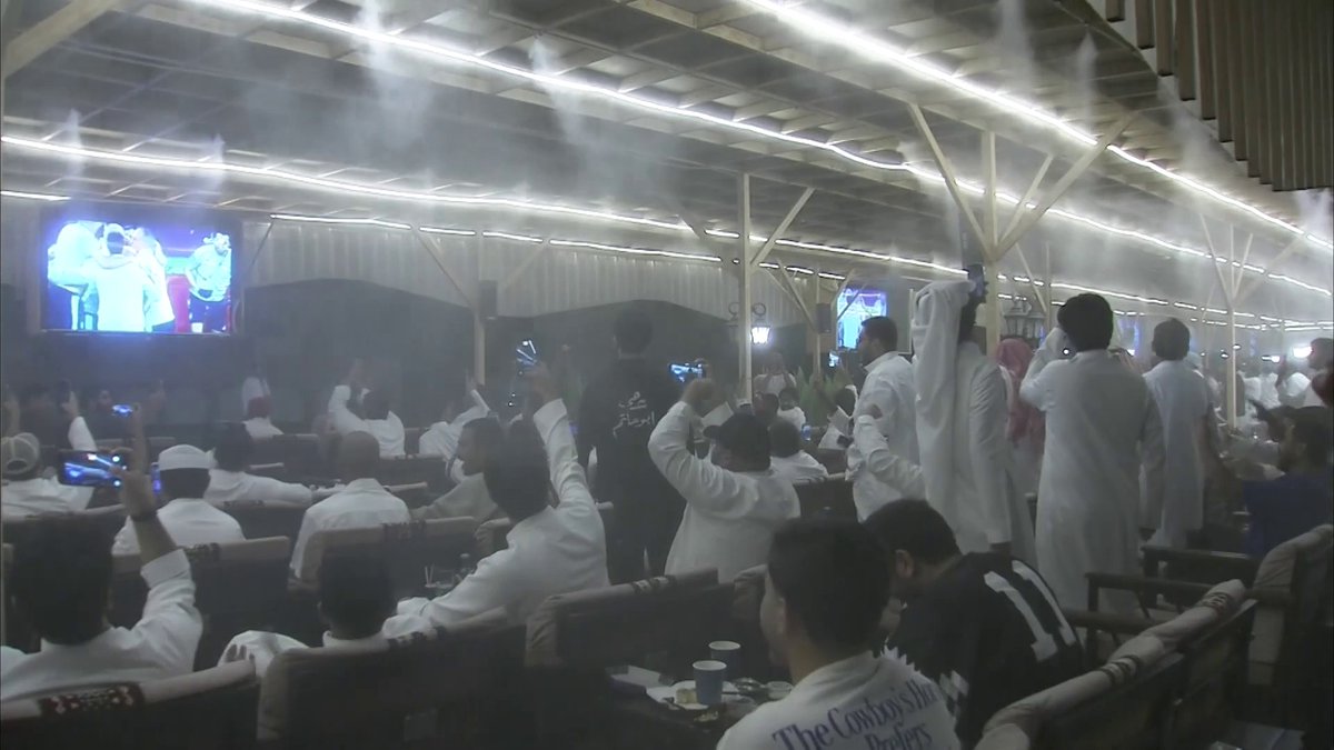 Fans wearing traditional white Saudi attire celebrate inside a mist-filled tent with visible text reading "The Cowboy Style" and "#Khaleeji_27," cheering as the Saudi national team wins the Gulf Cup final on large screens.