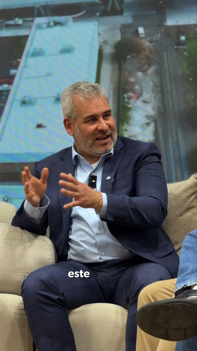 A man in a navy suit sits on a beige couch, gesturing animatedly with both hands while speaking; a screen behind him shows a city traffic scene, and the text "este" is overlaid near the bottom.