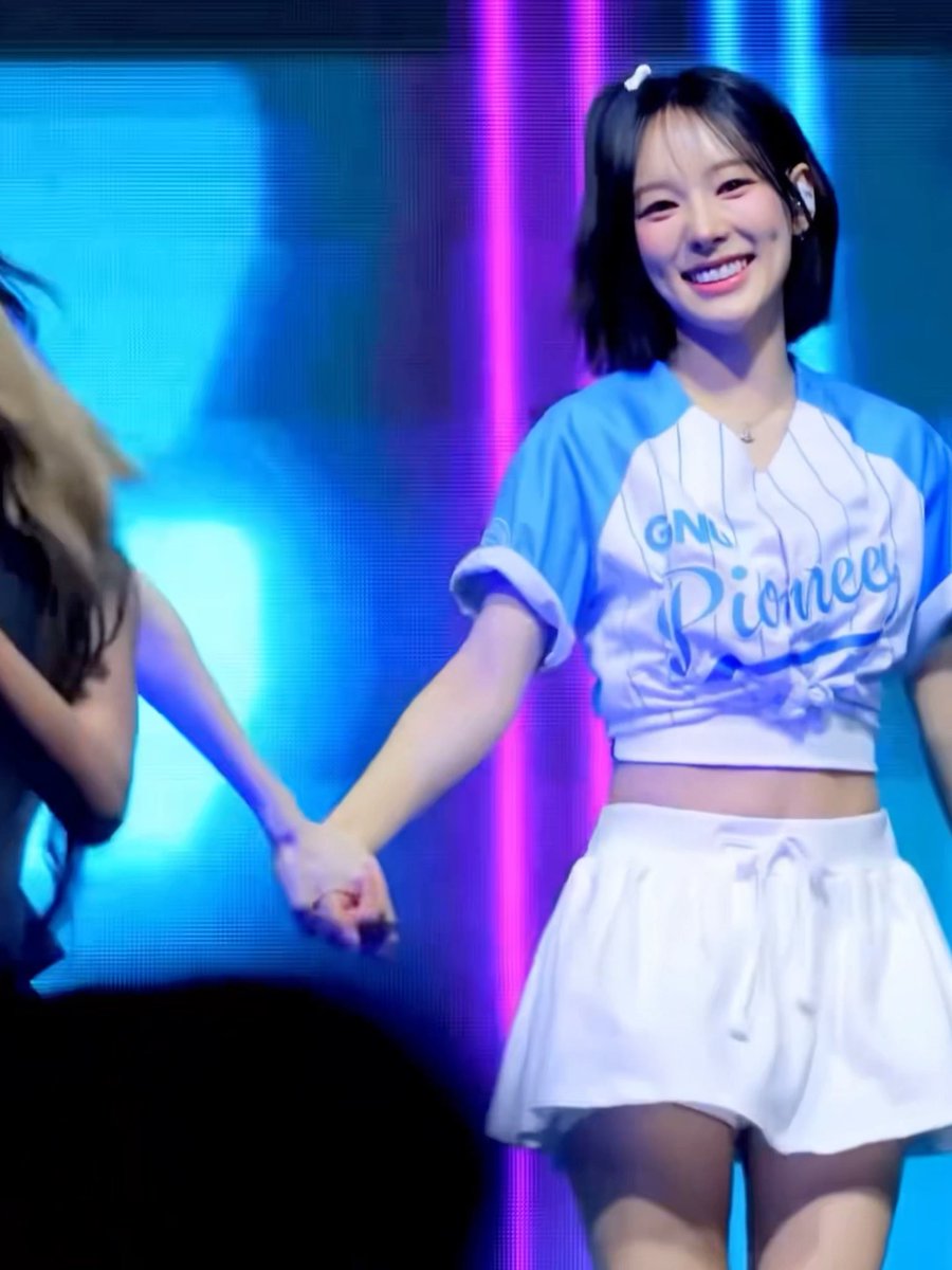A performer on stage is smiling brightly while wearing a blue and white baseball-style crop top that reads "GNUV Pioneers" along with a white pleated skirt, fitting the caption's reference to a "full senyum" moment.