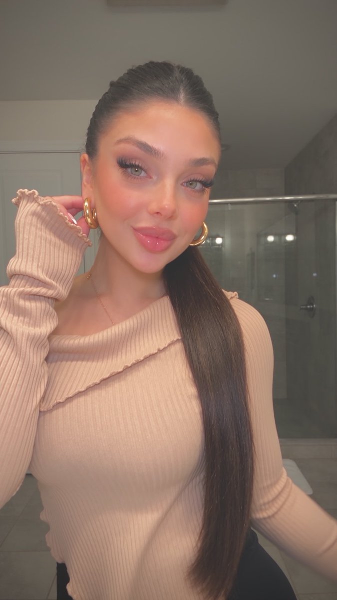 A young woman with long brunette hair in a sleek ponytail poses for a selfie, wearing a peach-colored ribbed off-shoulder top and gold hoop earrings, with flawless makeup featuring pink lips and defined lashes. The image perfectly embodies the "Nothing to somethin" caption, showcasing a polished, glamorous transformation that highlights a complete beauty and styling glow-up.