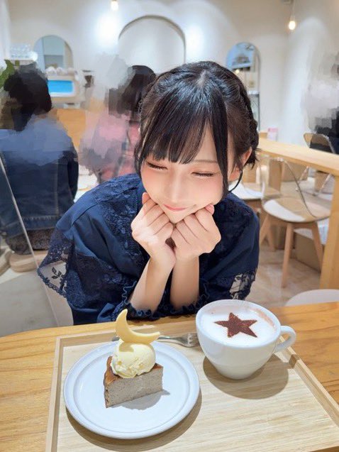 A smiling young woman with dark hair in a lace top posing with her chin on her hands at a cafe table, with a slice of cake topped with a moon and a hot drink with a star design in front of her.