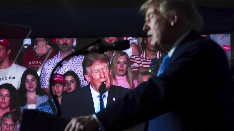 Donald Trump is shown on a large screen addressing a rally audience, with a man in a suit gesturing toward the display, likely during a televised event or broadcast. The image relates to the post by depicting the political figure central to the controversy surrounding his comments about potential Iranian strikes on US cities.