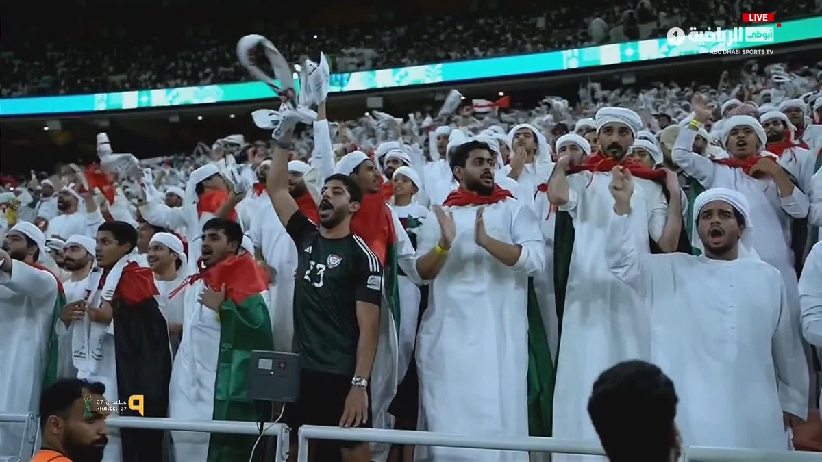 A passionate crowd of Emirati fans cheers in a stadium, dressed in traditional white kanduras and waving red, white, green, and black flags, with "UAE" and "#خليجي27" visible on screen, celebrating during the 27th Gulf Cup as highlighted in the post.