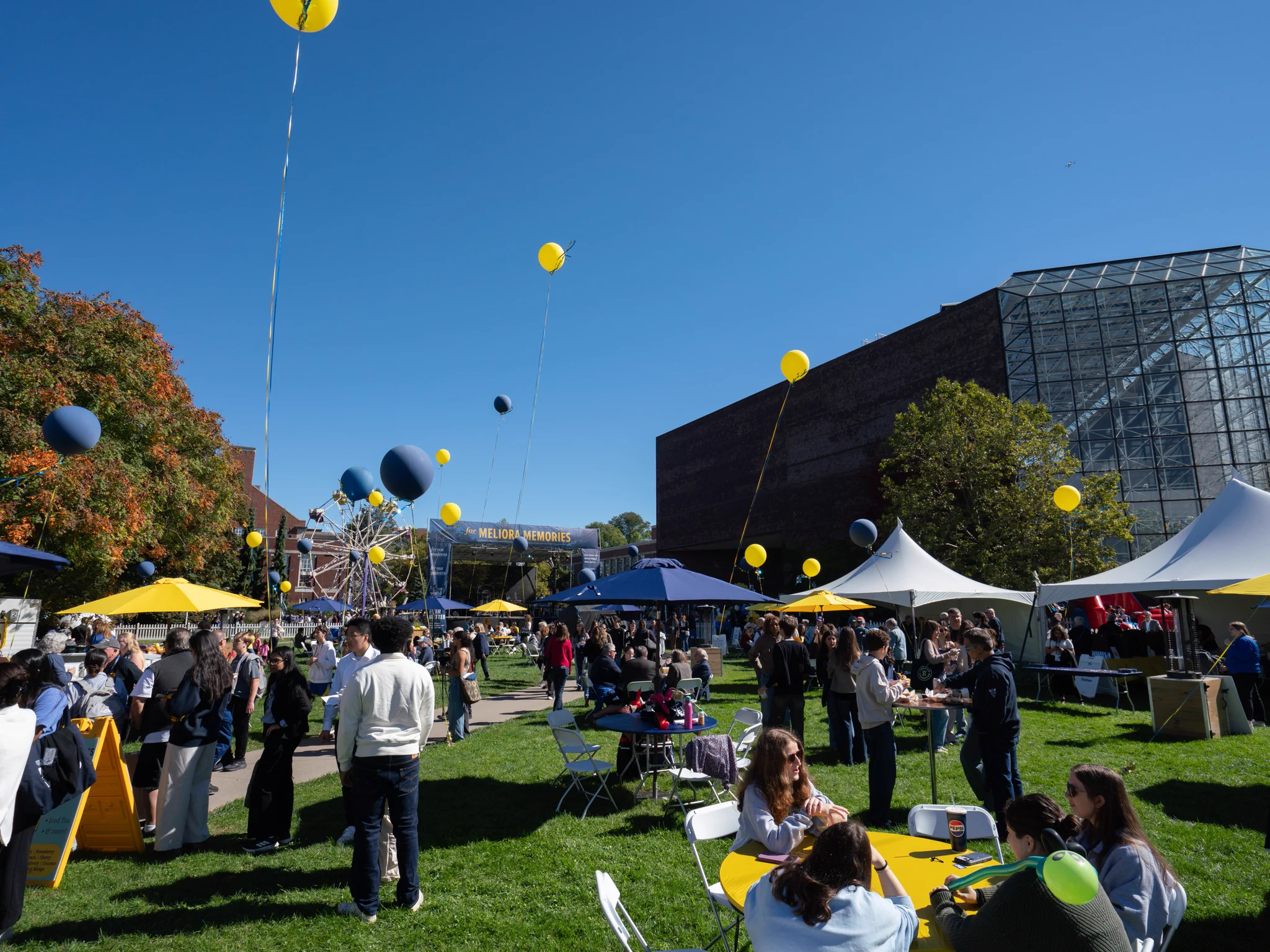 A vibrant outdoor event with people gathered at tables on a grassy area. Yellow and blue balloons are floating above, and there is a Ferris wheel in the background. Several tents and a large brick building with a glass facade are visible under a clear blue sky.