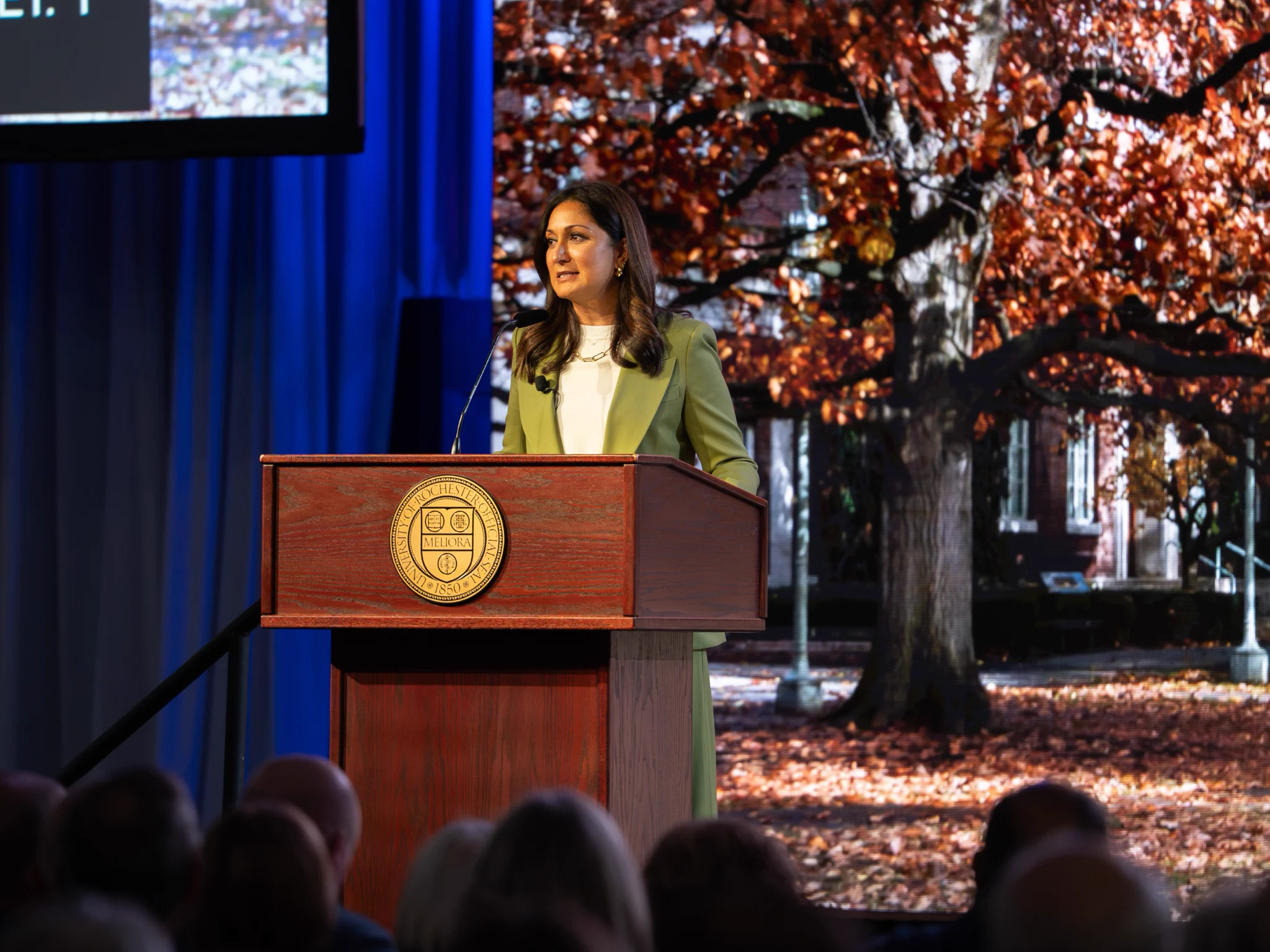 A person stands at a podium with a seal, giving a speech in front of an audience. The backdrop features a large tree with autumn leaves and a building partially visible.